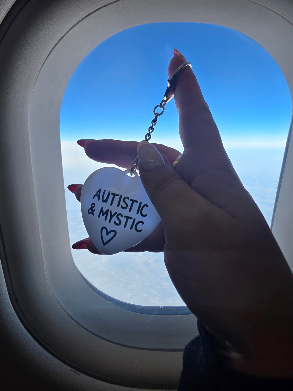Autistic & Mystic heart charm held in front of an airplane window above the clouds